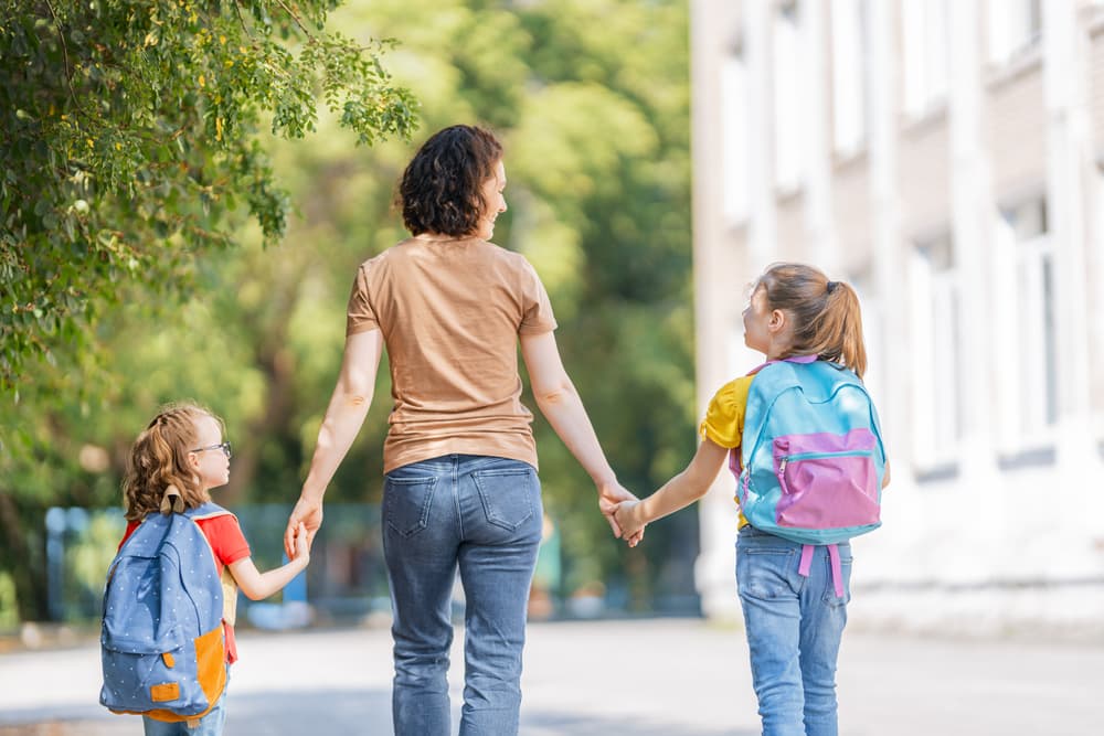 Parent and pupils of primary school go hand in hand. Woman and girls with backpacks behind backs. Beginning of lessons. First day of fall.
